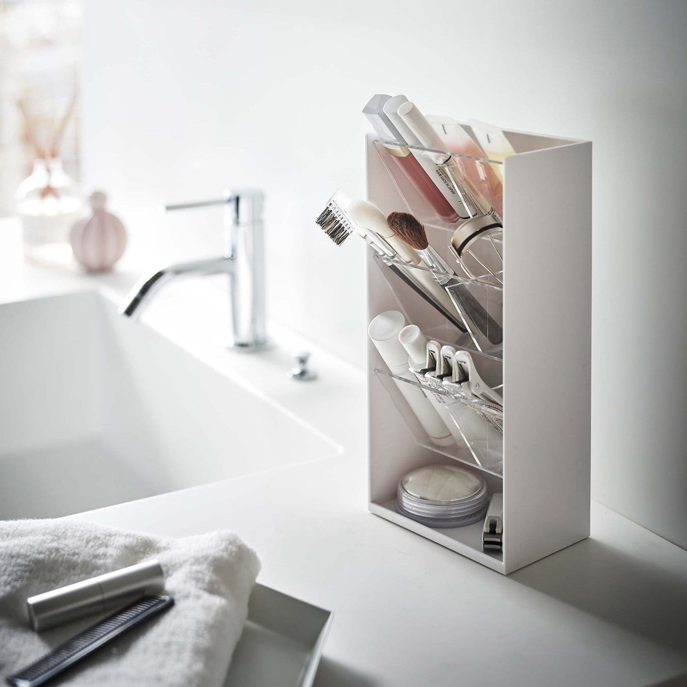 An angled view of a white rectangular resin cosmetics organizer on a white bathroom counter. It has an open face and top and three deep transparent trays that sit diagonally with adjustable transparent dividers placed in the middle of each tray for easy visibility.