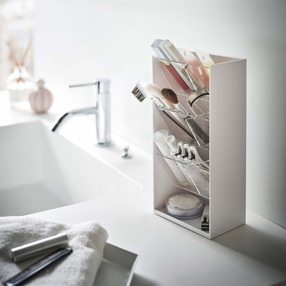 An angled view of a white rectangular resin cosmetics organizer on a white bathroom counter. It has an open face and top and three deep transparent trays that sit diagonally with adjustable transparent dividers placed in the middle of each tray for easy visibility.