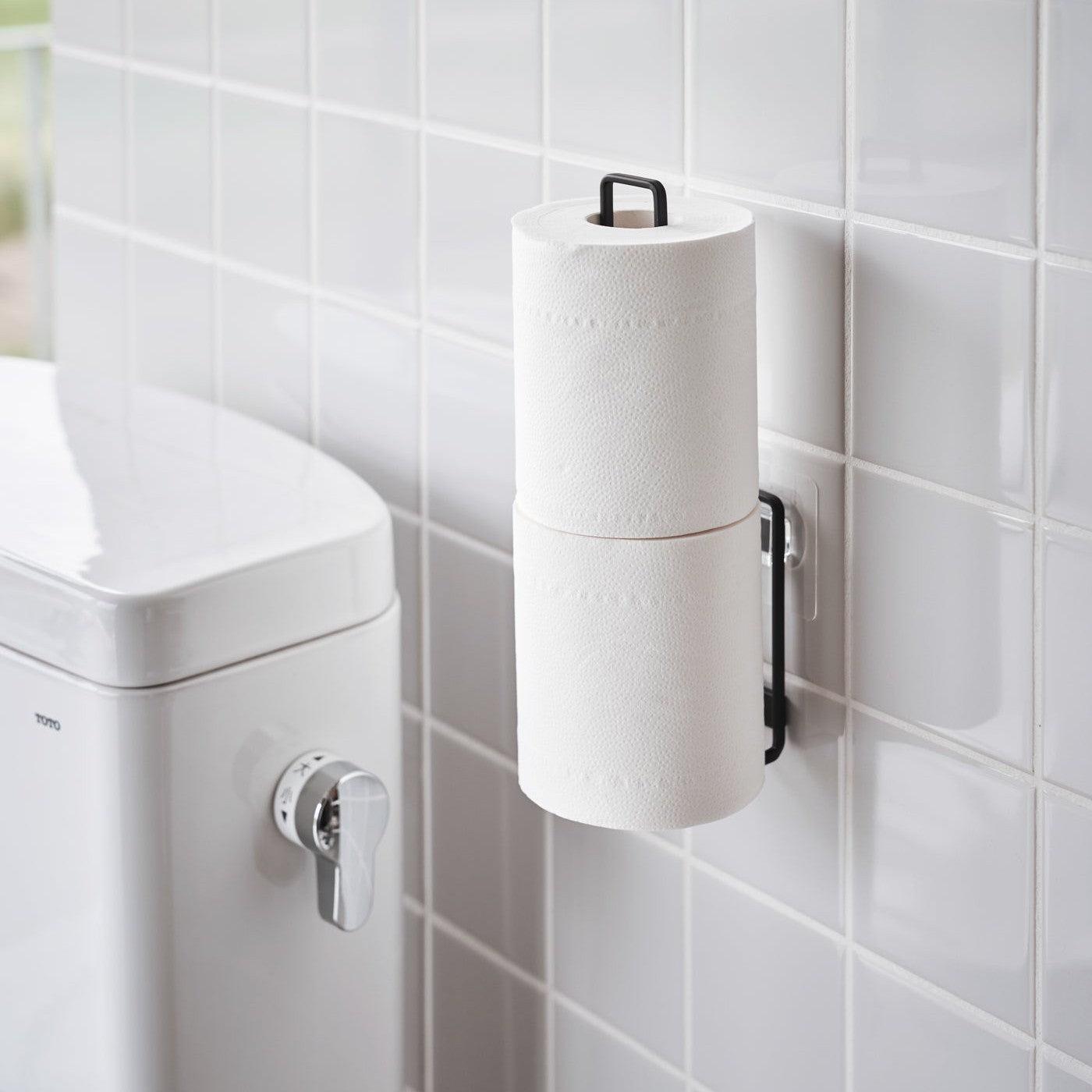 Black Yamazaki Home Traceless Adhesive Toilet Paper Holder attached to a wall with toilet paper rolls stored