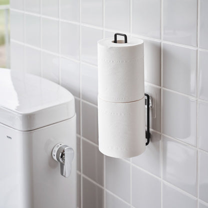 Black Yamazaki Home Traceless Adhesive Toilet Paper Holder attached to a wall with toilet paper rolls stored