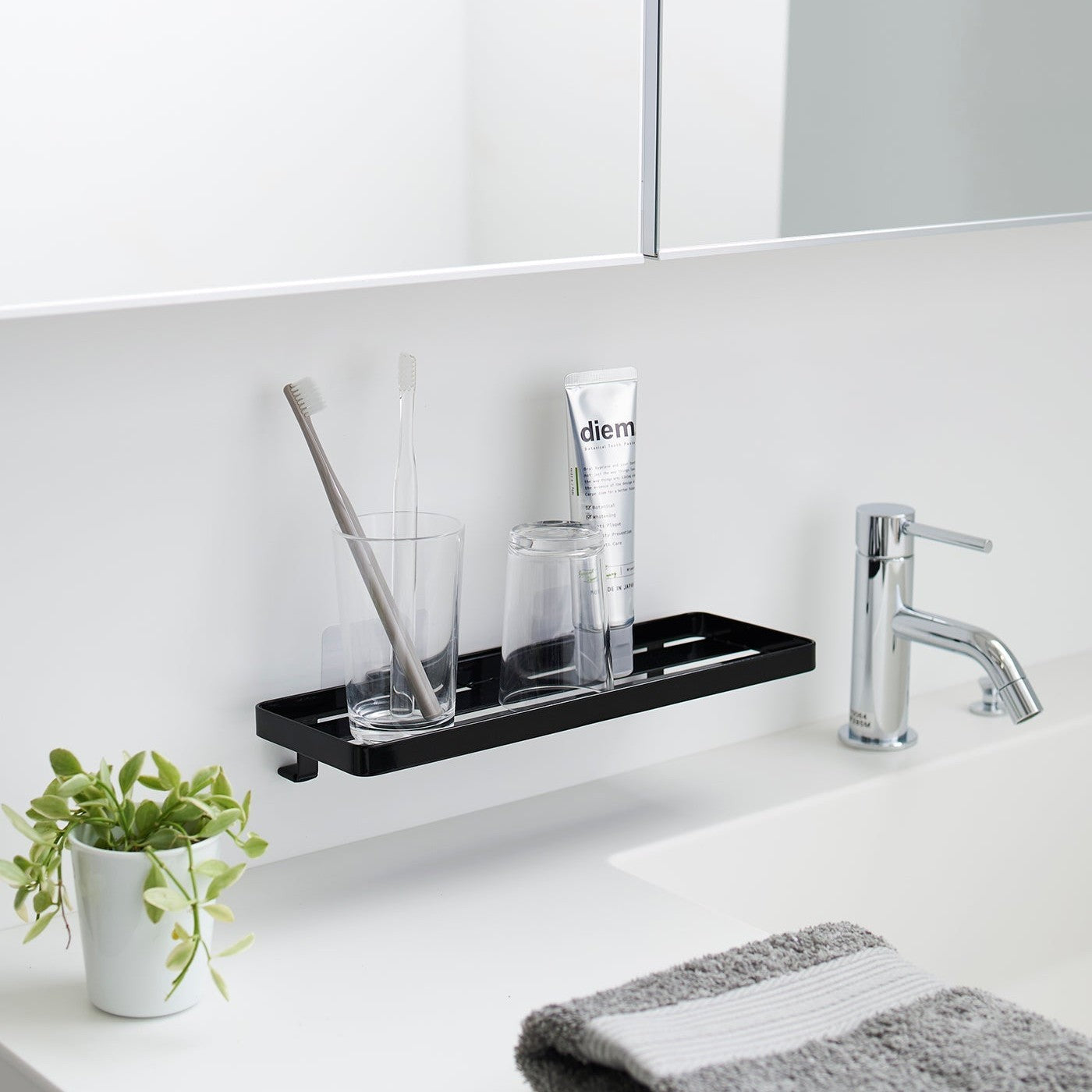 Black steel vanity tray is installed on a wall behind a sink via a translucent film hook. The tray has open slats for drainage and holds various skin care items.