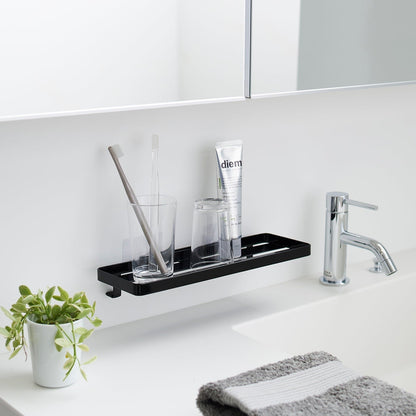 Black steel vanity tray is installed on a wall behind a sink via a translucent film hook. The tray has open slats for drainage and holds various skin care items.
