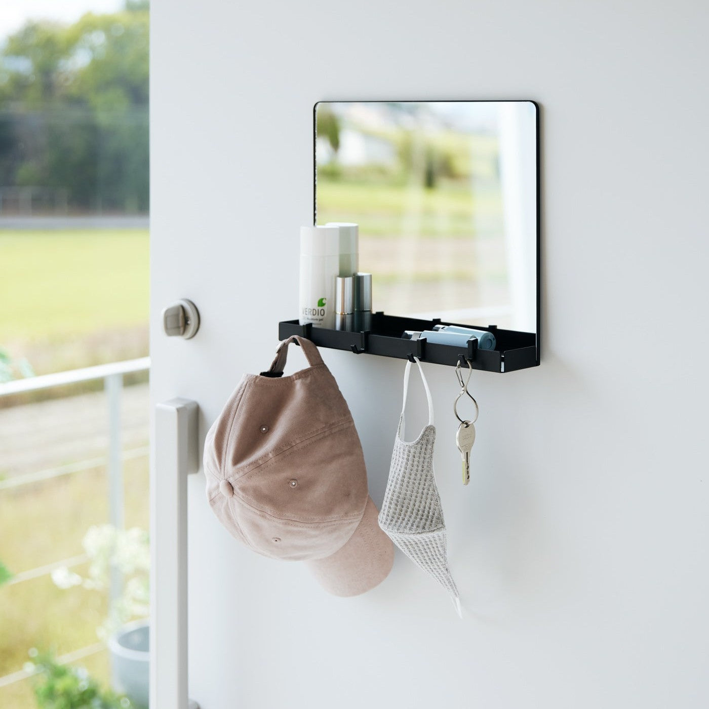 A black Yamazaki Magnetic Mirror With Storage Rack attached to a door with a hanging hat, mask, and key with a view outside.