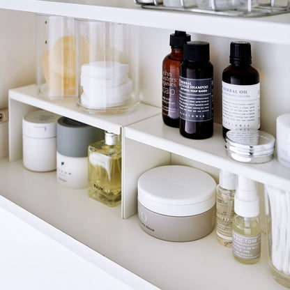 Open medicine cabinet showing two white countertop risers on bottom shelf, side by side. Upper level of the riser holds perfume and hair products, while skincare and small sprays are stored on the bottom.