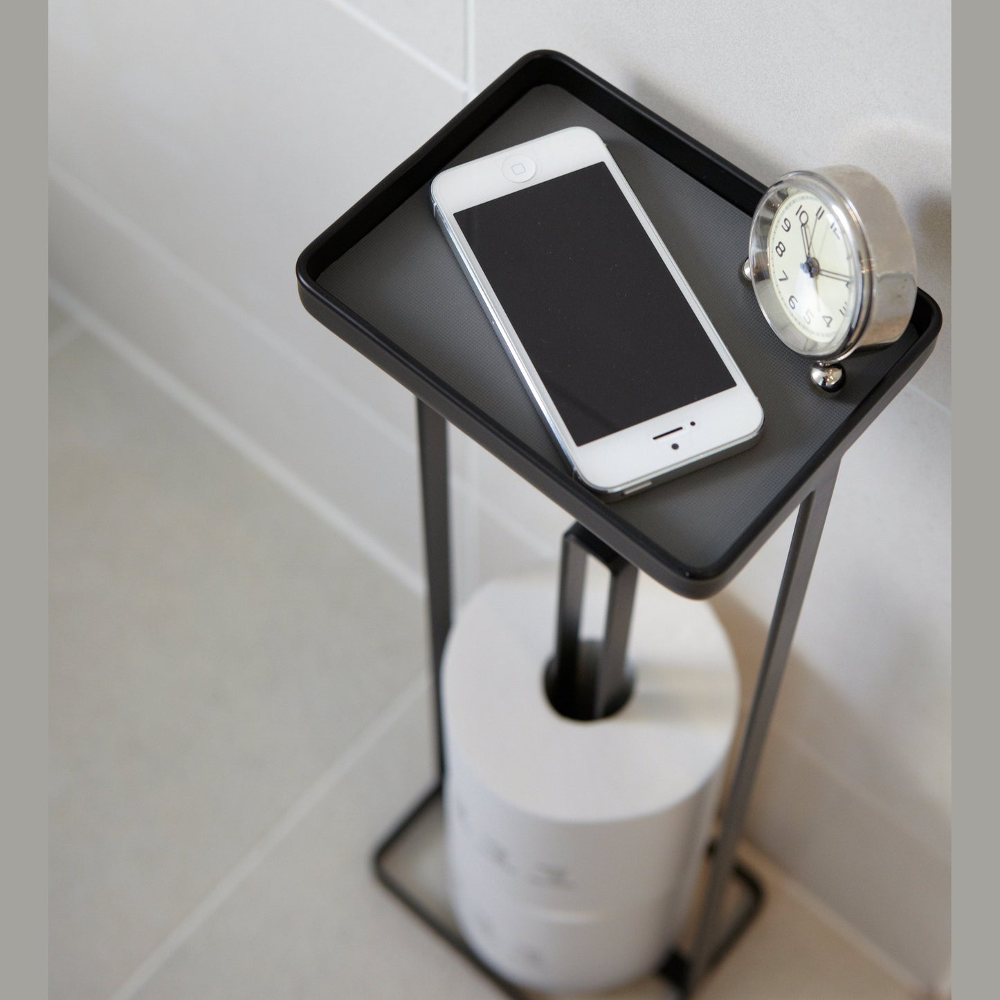 Close up aerial view of black Toilet Paper Stocker holding phone, clock, and toilet paper in bathroom by Yamazaki home.
