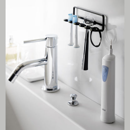 Yamazaki Home's black Traceless Adhesive Toothbrush Holder on a bathroom wall, holding two toothbrushes and a razor beside a faucet.