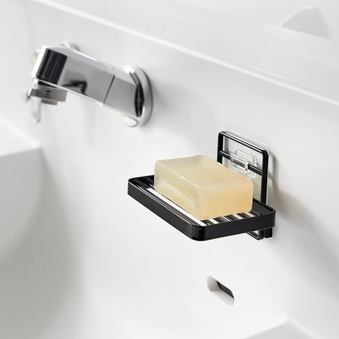 Yamazaki Home's black Traceless Adhesive Soap Tray mounted on a bathroom wall, holding a bar of yellow soap next to a chrome faucet.