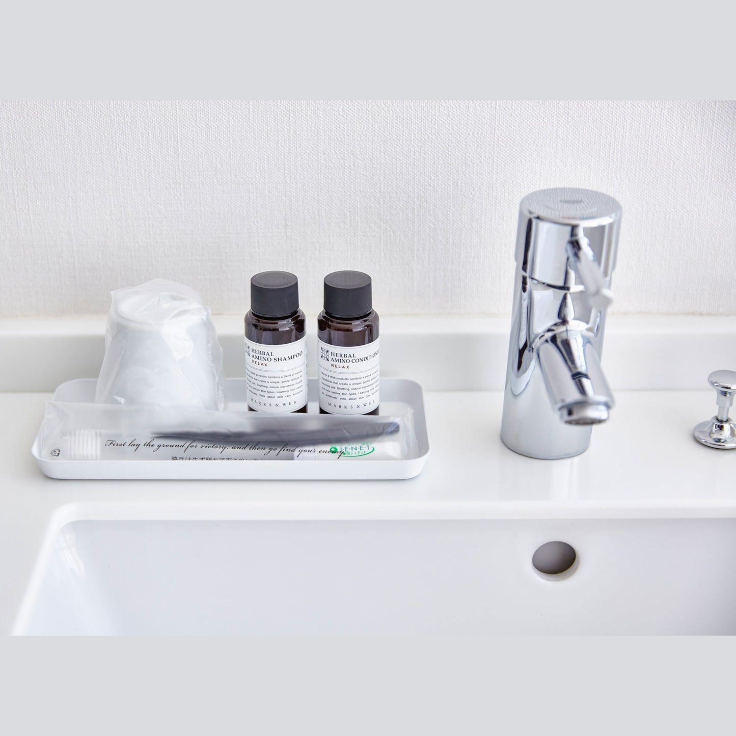 Front view of flat white Accessory Tray holding soap, cup, and toothbrush on sink countertop by Yamazaki Home.