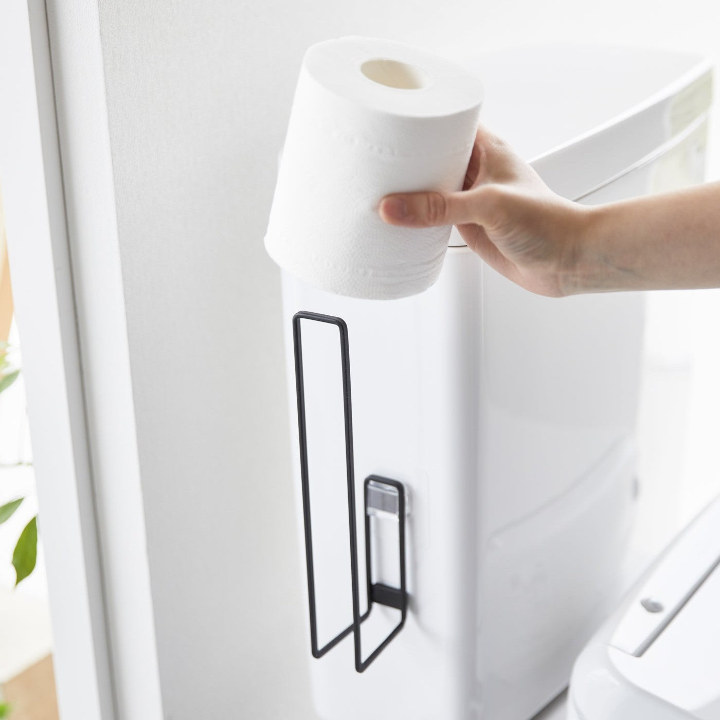 A toilet paper roll being placed on a black Yamazaki Home Traceless Adhesive Toilet Paper Holder