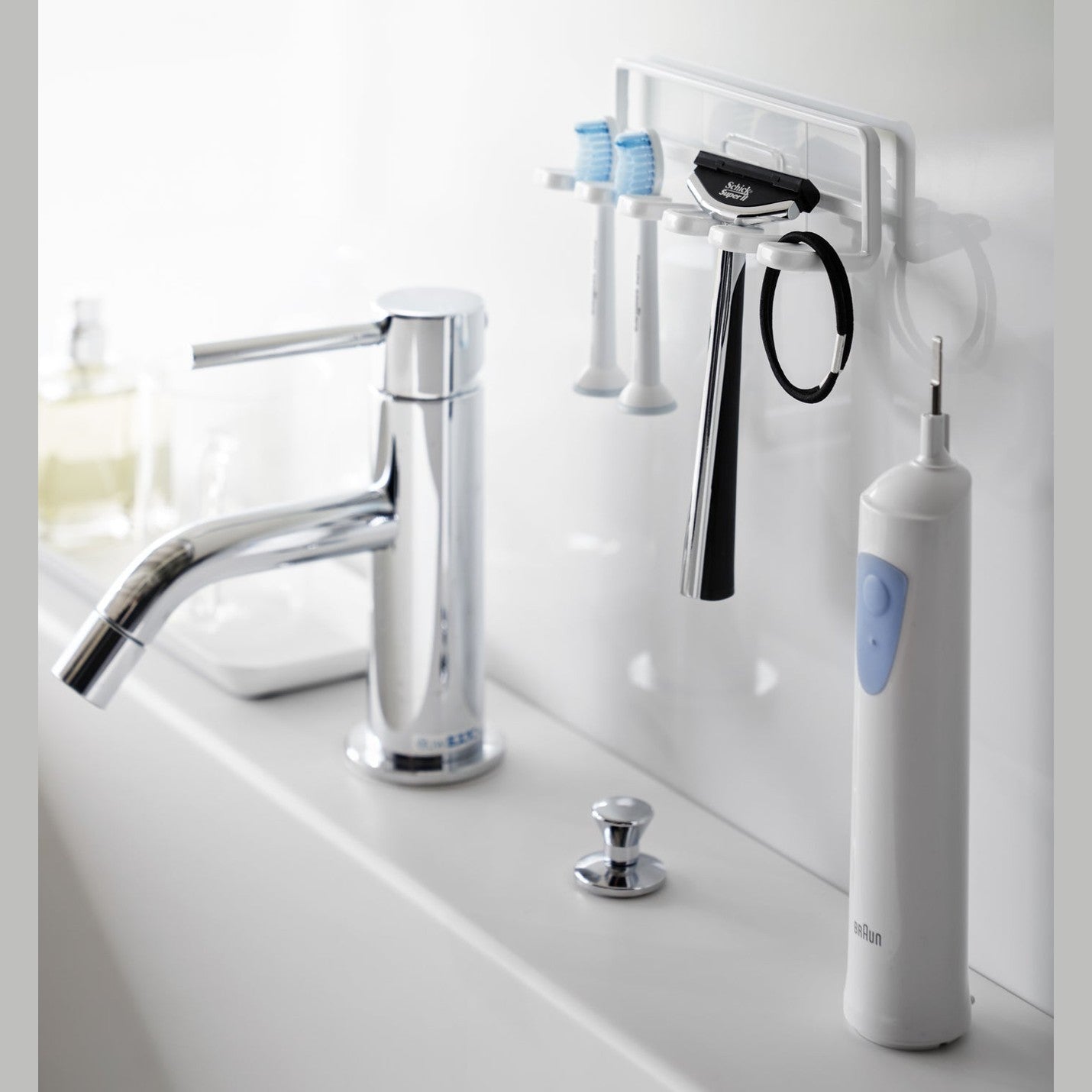 Yamazaki Home's white Traceless Adhesive Toothbrush Holder on a bathroom wall, holding two toothbrushes and a razor beside a faucet.
