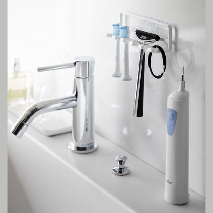 Yamazaki Home's white Traceless Adhesive Toothbrush Holder on a bathroom wall, holding two toothbrushes and a razor beside a faucet.