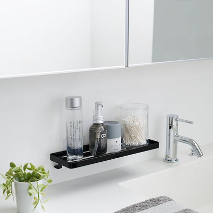 Black steel vanity tray is installed on a wall behind a sink via a translucent film hook. The tray has open slats for drainage and holds various skin care items.