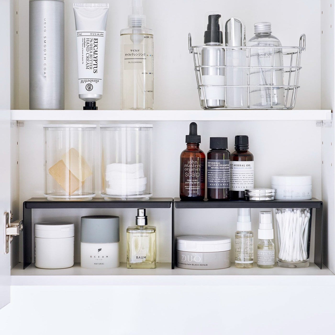 Open medicine cabinet showing two black countertop risers on bottom shelf, side by side. Upper level of the riser holds perfume and hair products, while skincare and small sprays are stored on the bottom.