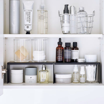 Open medicine cabinet showing two black countertop risers on bottom shelf, side by side. Upper level of the riser holds perfume and hair products, while skincare and small sprays are stored on the bottom.