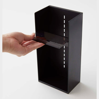 A male hand pulls a black transparent tray to adjust the location in a cosmetics organizer. It is a black resin rectangular cosmetics holder with an open face and top. The removable tray acts as a shelf and has an upward facing lip along the edge to prevent products from falling out.