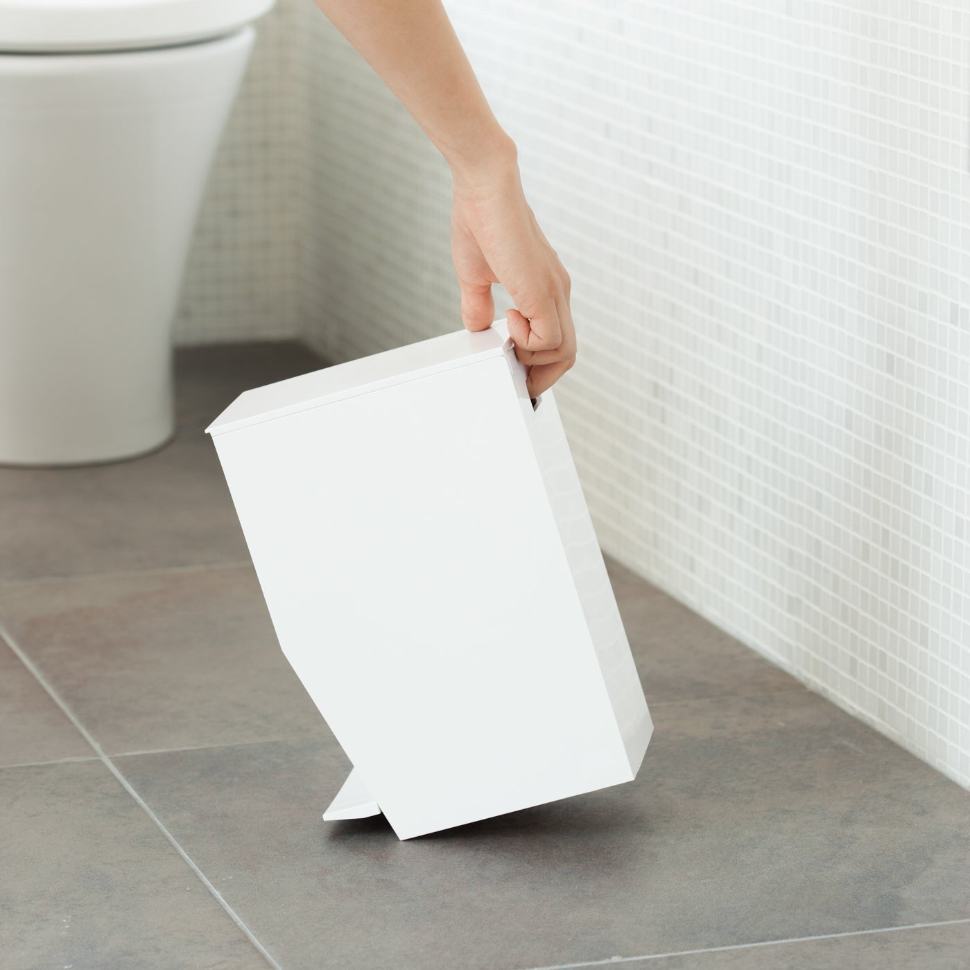 Side view of white Step Trash Can held by handle in bathroom by Yamazaki Home.