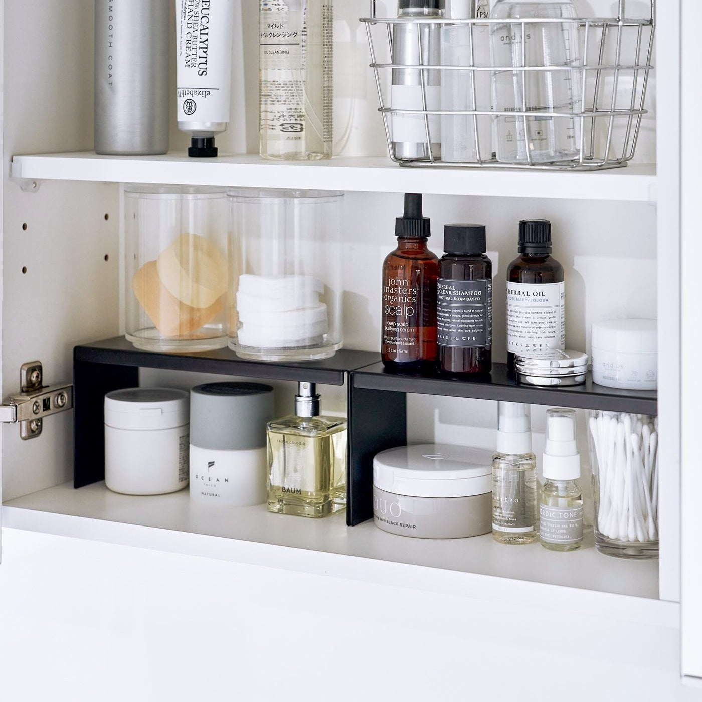 Open medicine cabinet showing two black countertop risers on bottom shelf, side by side. Upper level of the riser holds perfume and hair products, while skincare and small sprays are stored on the bottom.