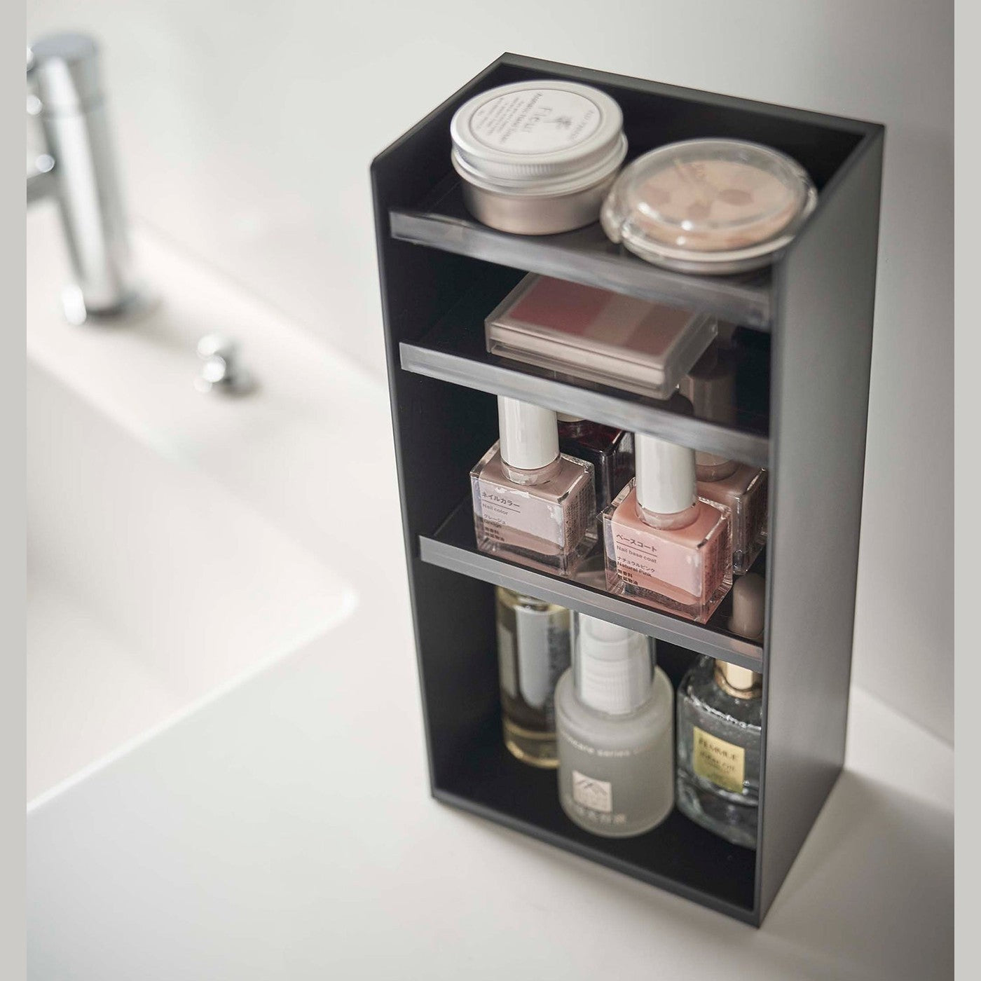 Angled view of a black rectangular cosmetics organizer sitting on a white bathroom counter. The organizer has three black transparent shelves with upward facing lips to prevent the products from falling-out.