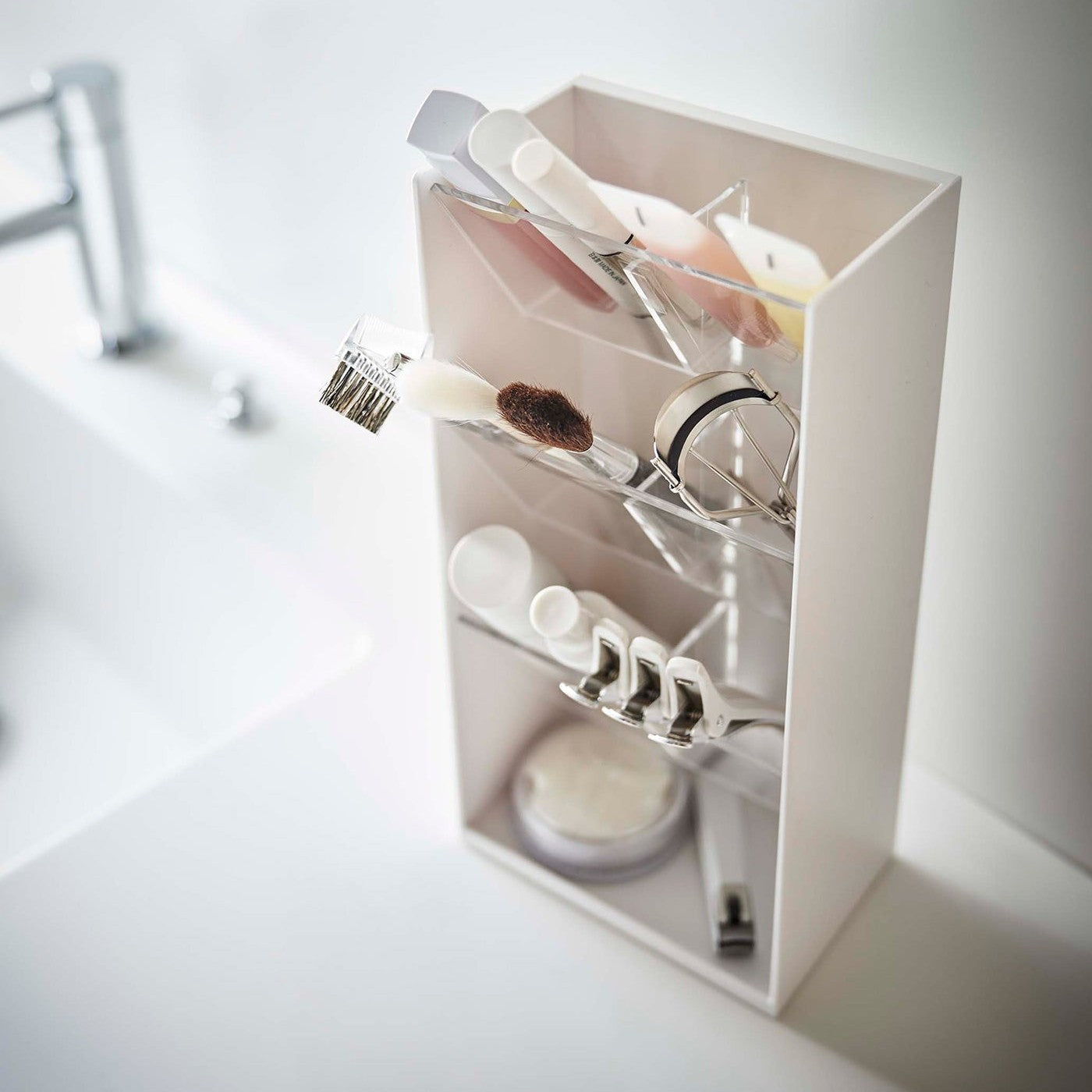 An angled view of a white rectangular resin cosmetics organizer on a white bathroom counter. It has an open face and top and three deep transparent trays that sit diagonally with adjustable transparent dividers placed in the middle of each tray for easy visibility.
