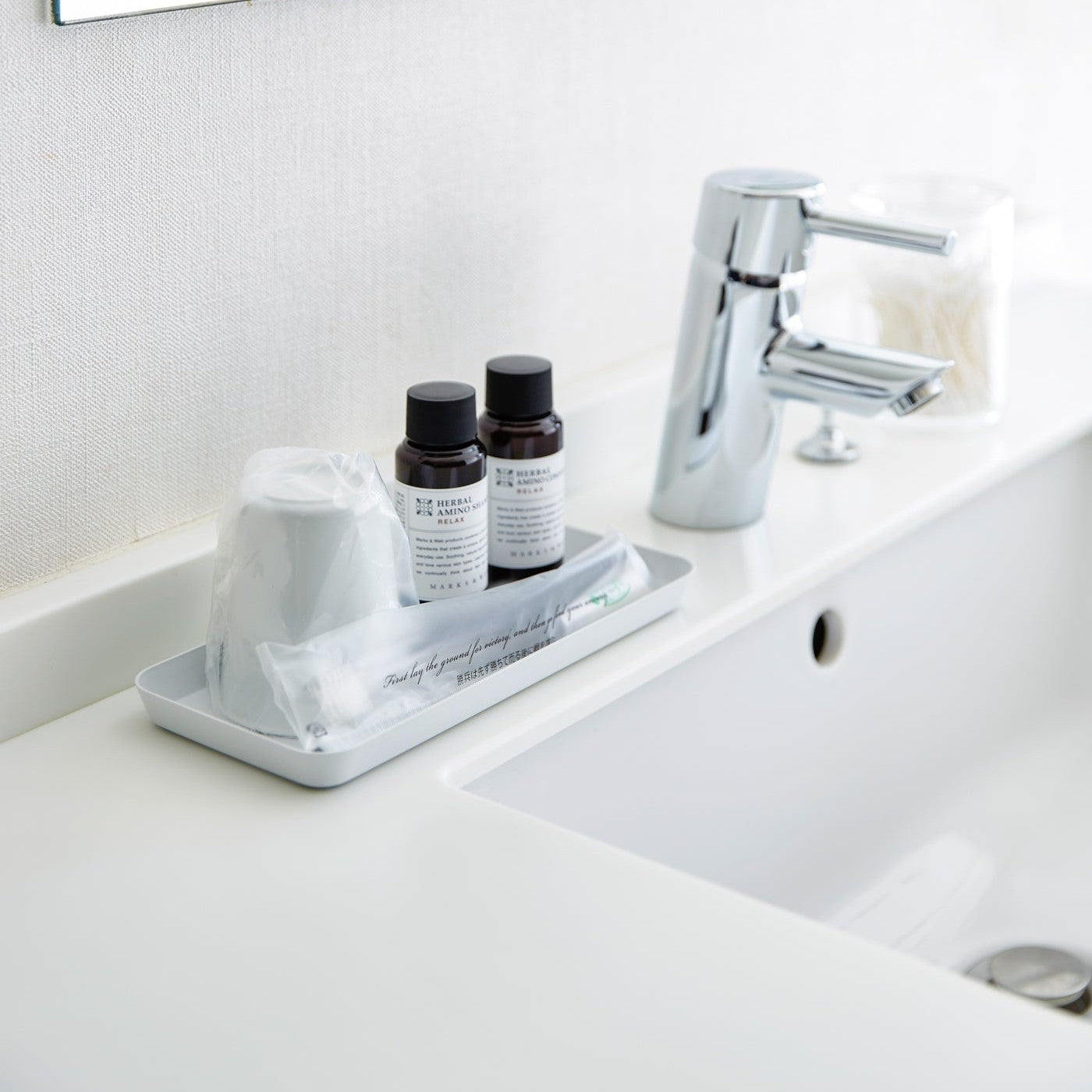 Flat white Accessory Tray holding soap, cup, and toothbrush on bathroom sink countertop by Yamazaki Home.