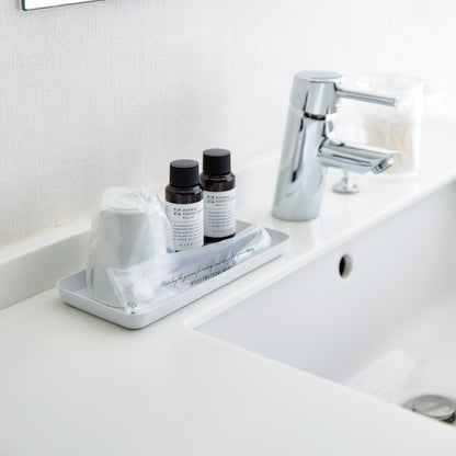 Flat white Accessory Tray holding soap, cup, and toothbrush on bathroom sink countertop by Yamazaki Home.