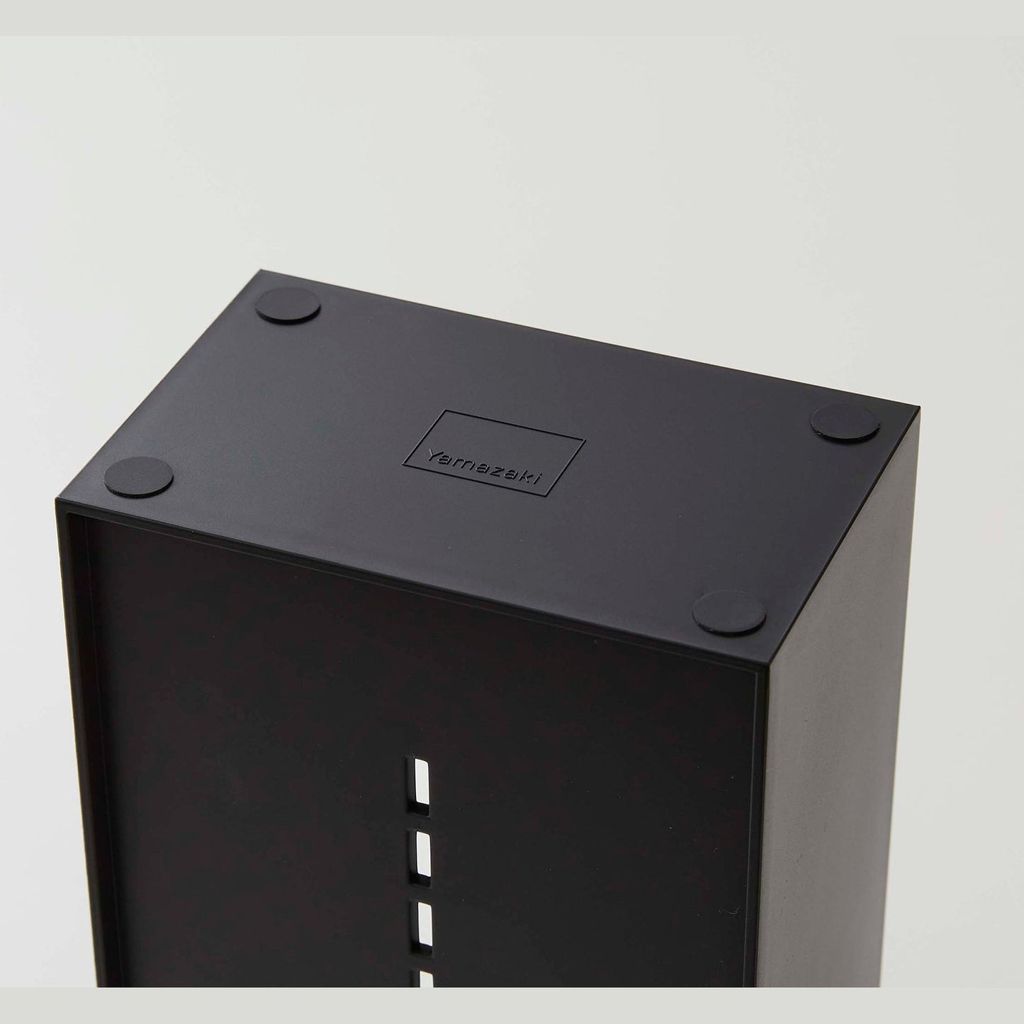 The underside of a black cosmetics organizer is shown. Four circular silicone rubber adhesives are on each of the corners to prevent sliding or surface damage. In the middle of the product is an engraving of the â€˜Yamazakiâ€™ logo, a rectangle with â€˜Yamazakiâ€™ printed in the bottom center.