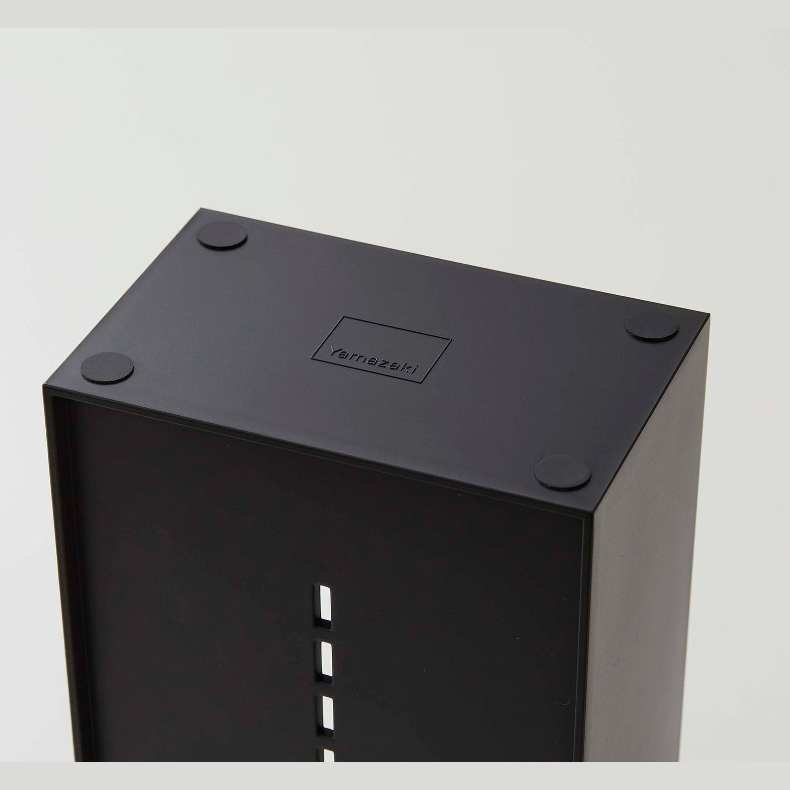 The underside of a black cosmetics organizer is shown. Four circular silicone rubber adhesives are on each of the corners to prevent sliding or surface damage. In the middle of the product is an engraving of the â€˜Yamazakiâ€™ logo, a rectangle with â€˜Yamazakiâ€™ printed in the bottom center.
