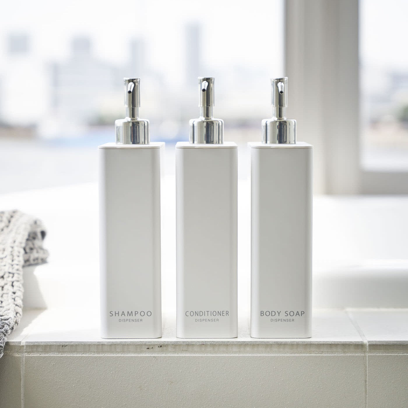Front view of Yamazaki Home white Shampoo, Conditioner, and Body Soap dispensers on the ledge of a modern bathtub.