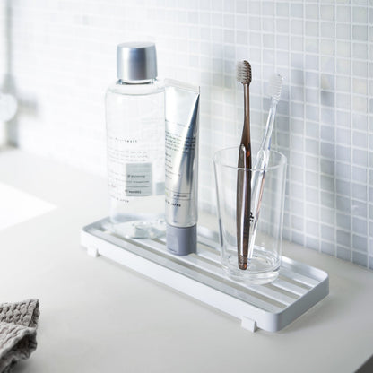 White slotted tray holding toothbrush and mouthcare items on bathroom sink counter by Yamazaki Home.