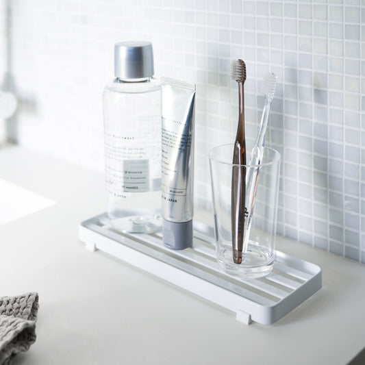 White slotted tray holding toothbrush and mouthcare items on bathroom sink counter by Yamazaki Home.