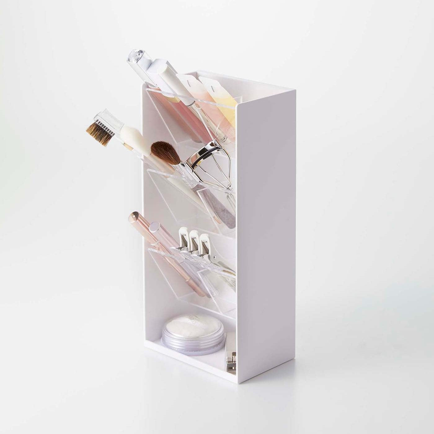 On top of a white surface is a white resin rectangular cosmetics organizer with an open face and top. It has three deep transparent trays that sit diagonally with adjustable transparent dividers placed in the middle of each tray for easy visibility. The bottom of the organizer has a slightly raised surface and acts as additional storage. Various cosmetics are stored in each tray and the bottom of the organizer holds nail clippers and a circular cosmetic product.