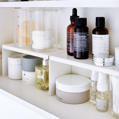 Open medicine cabinet showing two white countertop risers on bottom shelf, side by side. Upper level of the riser holds perfume and hair products, while skincare and small sprays are stored on the bottom.