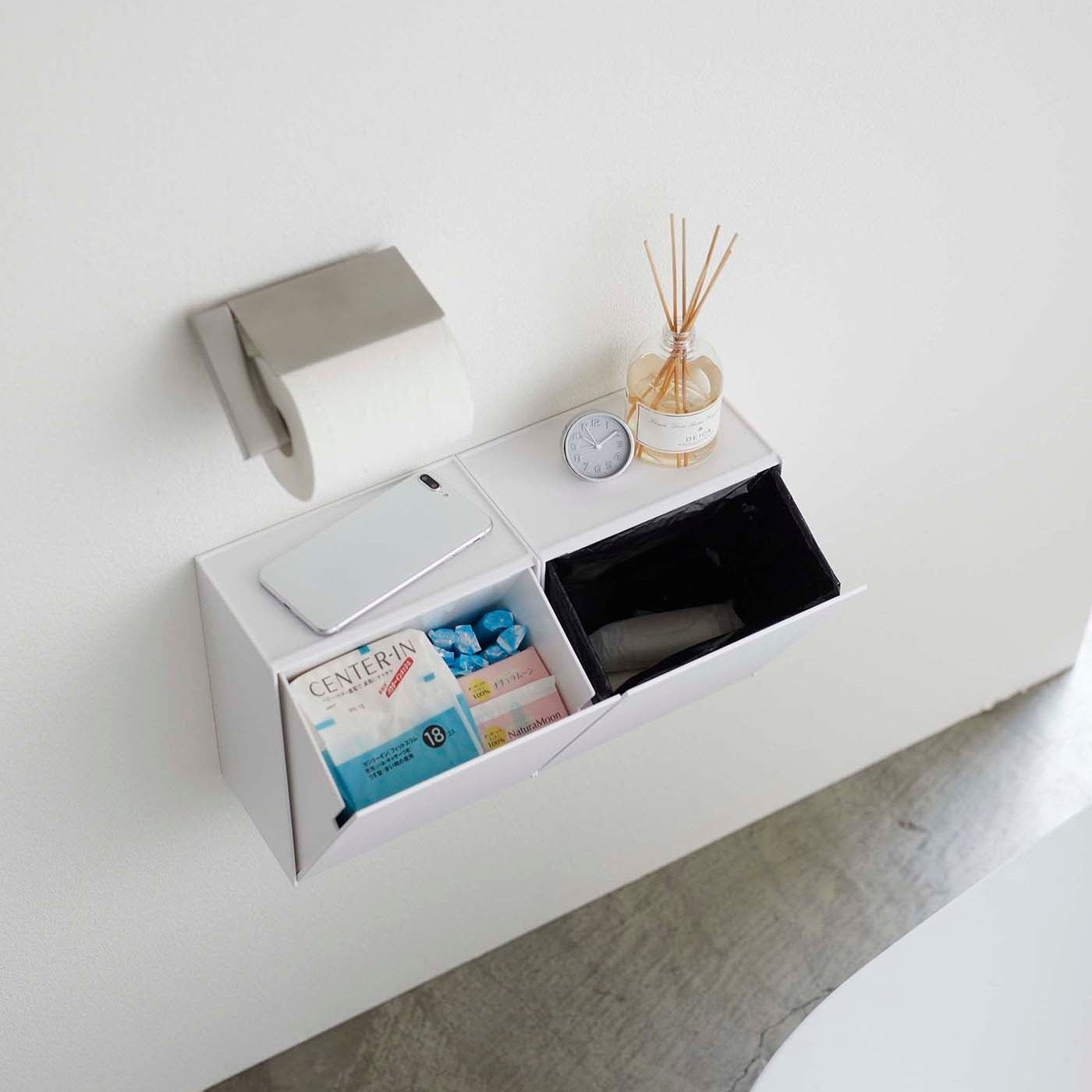 Aerial view of two open white Wall-Mount Storage bins holding items and trash bag in bathroom by Yamazaki Home.