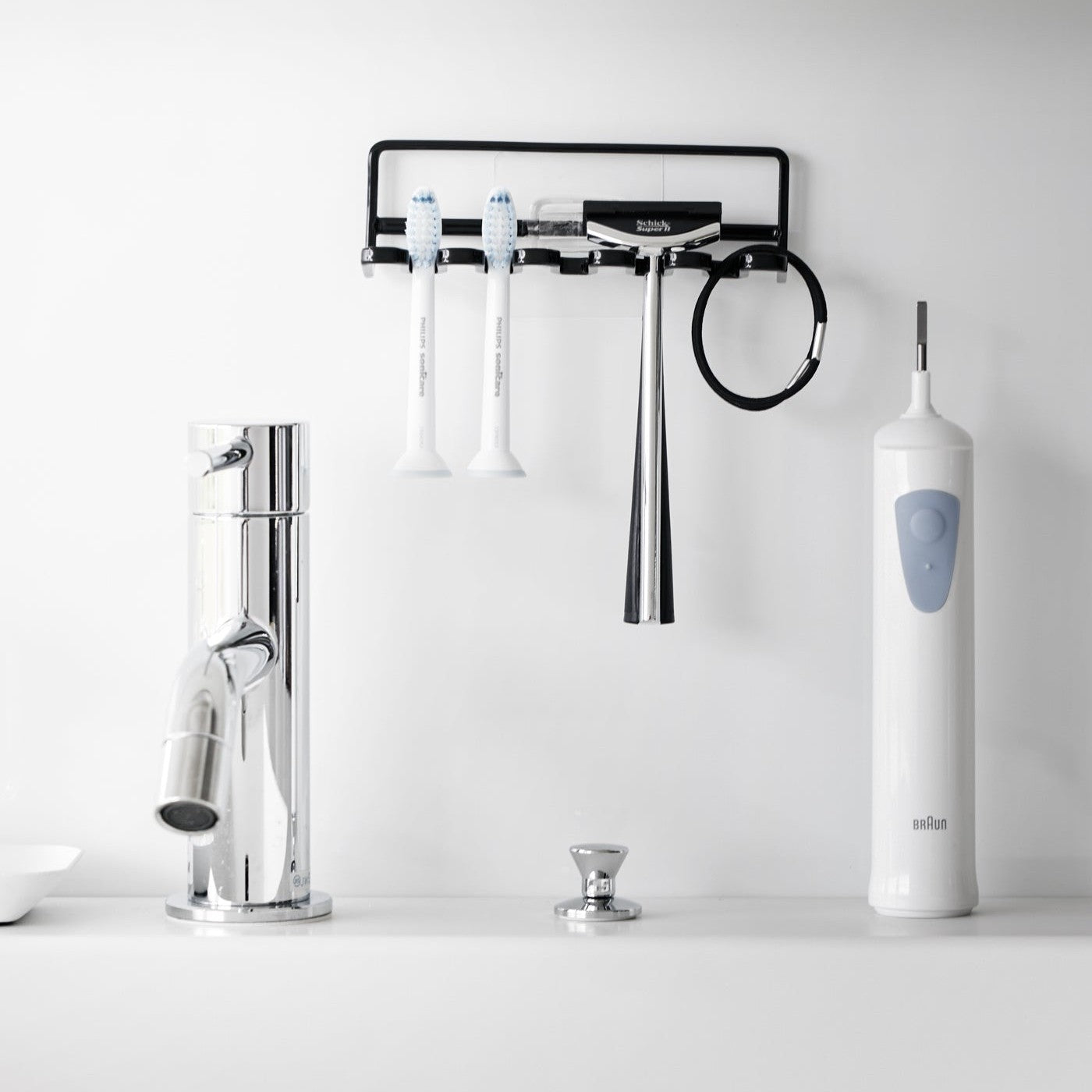 Yamazaki Home's black Traceless Adhesive Toothbrush Holder on a bathroom wall, holding two toothbrushes and a razor beside a faucet.