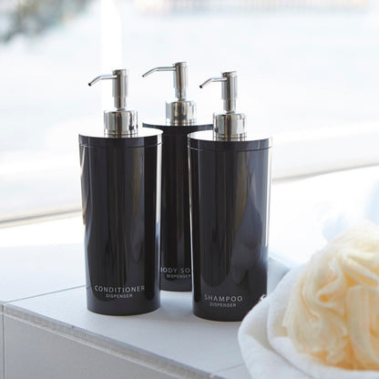 Yamazaki Home black Shampoo, Conditioner, and Body Soap dispensers next to a loofa and towel in bathroom.