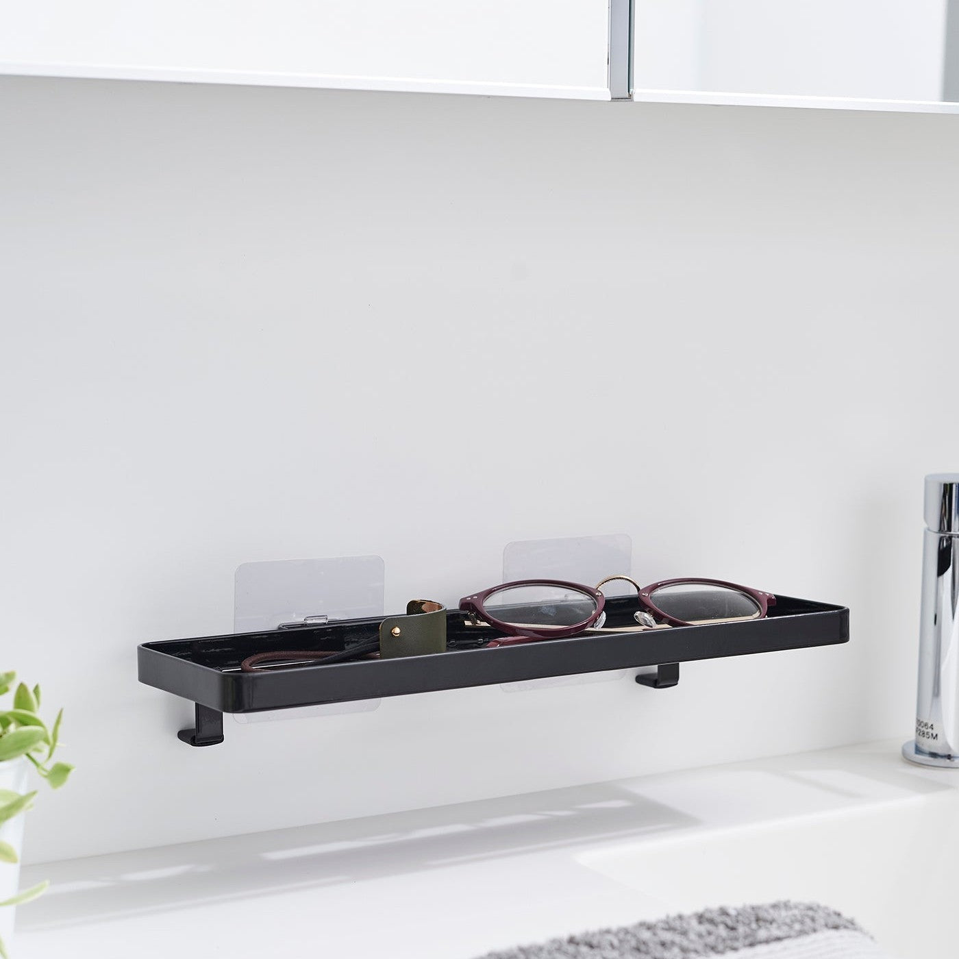 Black steel vanity tray is installed on a wall behind a sink via a translucent film hook. The tray has open slats for drainage and holds hair ties and reading glasses.