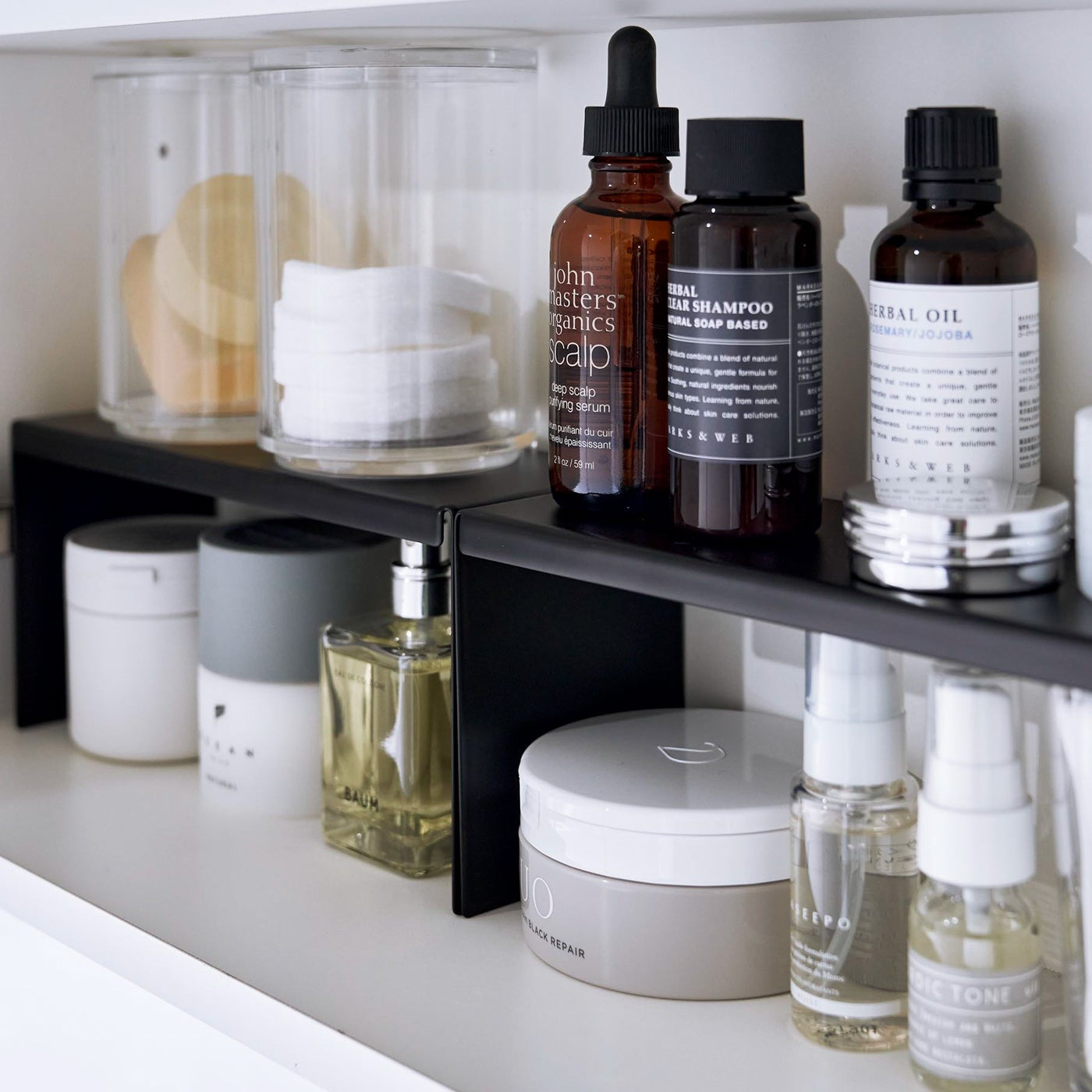 Open medicine cabinet showing two black countertop risers on bottom shelf, side by side. Upper level of the riser holds perfume and hair products, while skincare and small sprays are stored on the bottom.