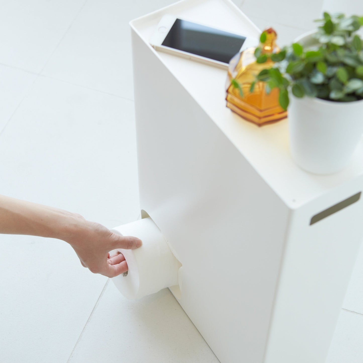 Aerial view of white Toilet Paper Stocker displaying phone, jar, and plant on top and dispensing a toilet paper roll by Yamazaki Home.
