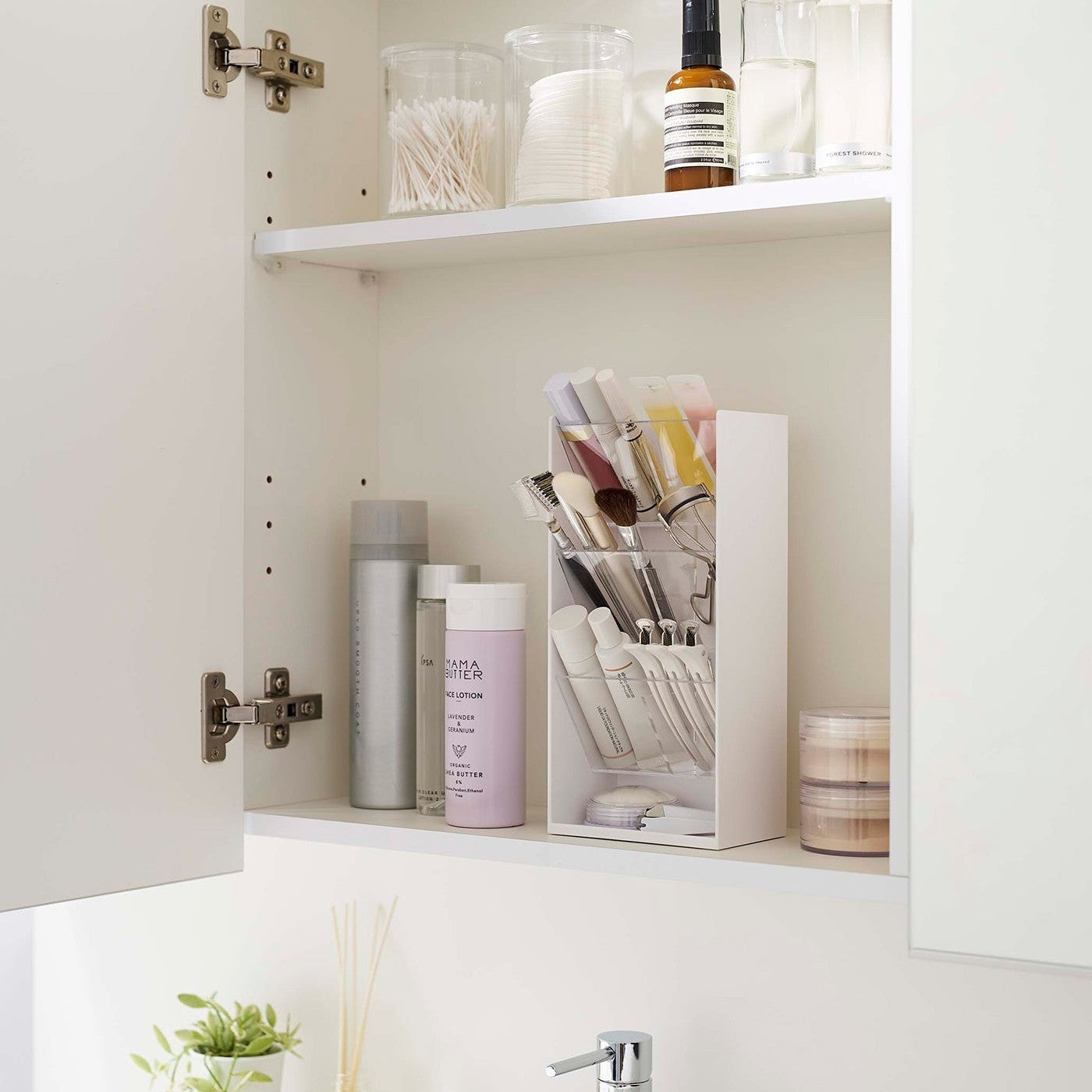 An angled view of an opened white medicine cabinet. Sunlight is focused on the right upper corner. The top half of the cabinet holds face serums and two clear containers with cotton swabs and rounds.