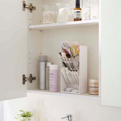 An angled view of an opened white medicine cabinet. Sunlight is focused on the right upper corner. The top half of the cabinet holds face serums and two clear containers with cotton swabs and rounds.